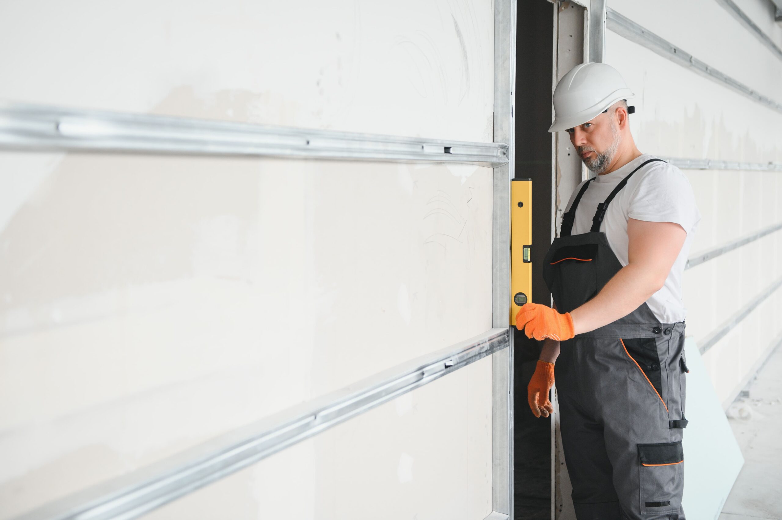 Construction Worker Ensuring Drywall Level on Metal Framing Construction worker wearing a white hard hat, white T-shirt, gray overalls with orange trim, and orange gloves, carefully checking the vertical alignment of a newly installed drywall panel against a metal stud framing using a yellow spirit level.