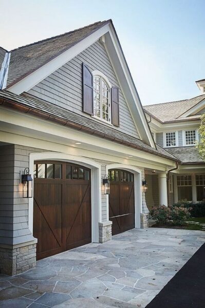 Exterior of a traditional home featuring two dark-stained wood carriage house style garage doors with arched windows, set into a facade of light grey shingle siding.